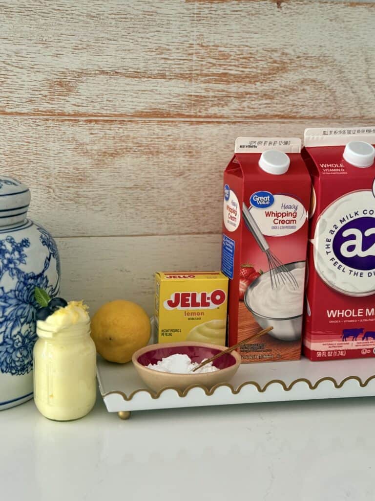 Jell-O lemon instant pudding mix, milk, heavy whipping cream, lemon powdered sugar in a bowl all on a tray and beside it is a blue and white flowered vase with simple lemon pudding dessert in a small glass jar all on the countertop