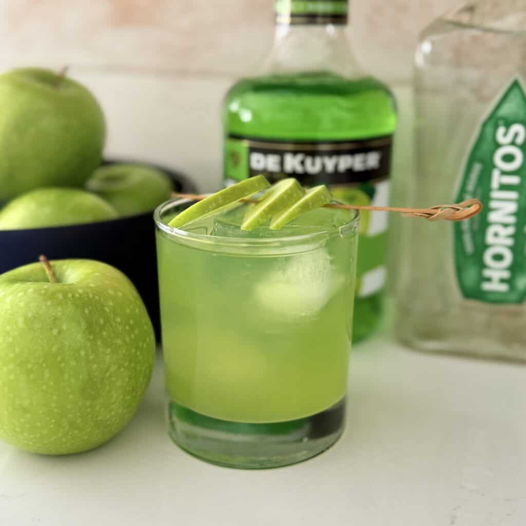 The sour apple margarita is on the counter with green apple slices on top of green apples in a black bowl behind it, a green apple beside it, a bottle of sour apple schnapps behind, and a bottle of white tequila all on the counter.