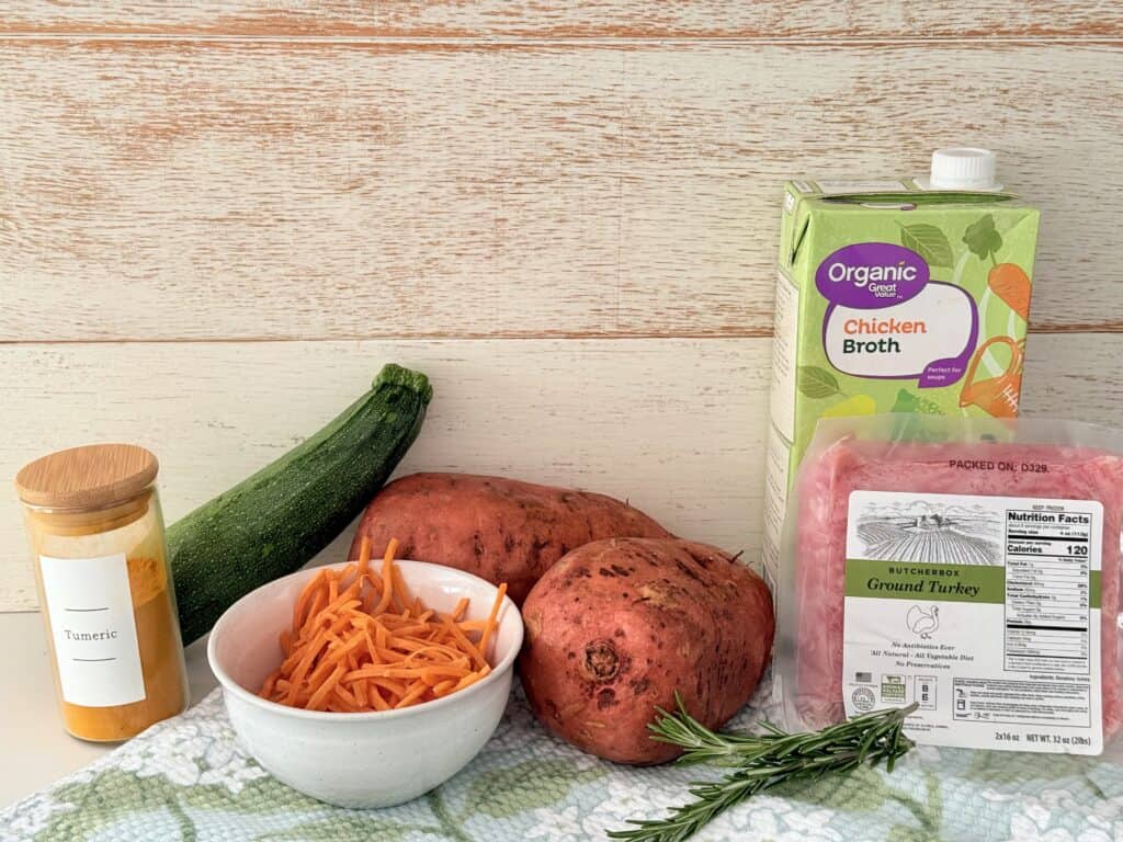 Ground turkey meat, chicken, broth, two medium size, sweet potatoes, shredded carrots, and a white bowl two sprigs of rosemary, a zucchini and turmeric seasoning all on a flower print cloth on the counter