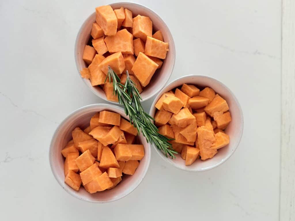 Chopped up sweet potatoes and bite-size pieces in three separate white bowls on the counter with two sprigs of rosemary on top of them