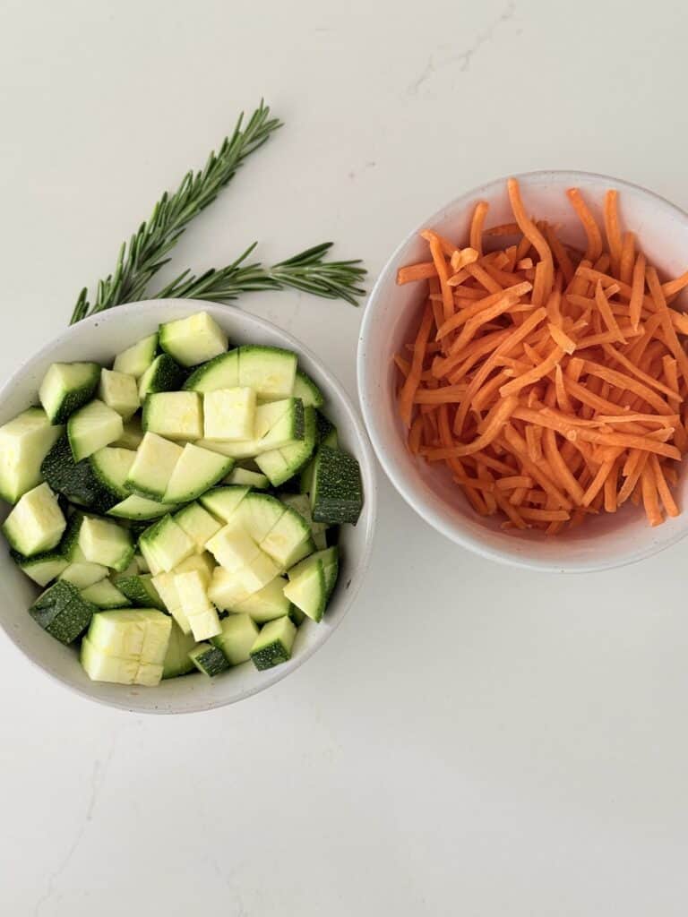 Diced zucchini in a bowl on the counter and shredded carrots in a bowl on the counter with two sprigs of rosemary behind them on the counter.