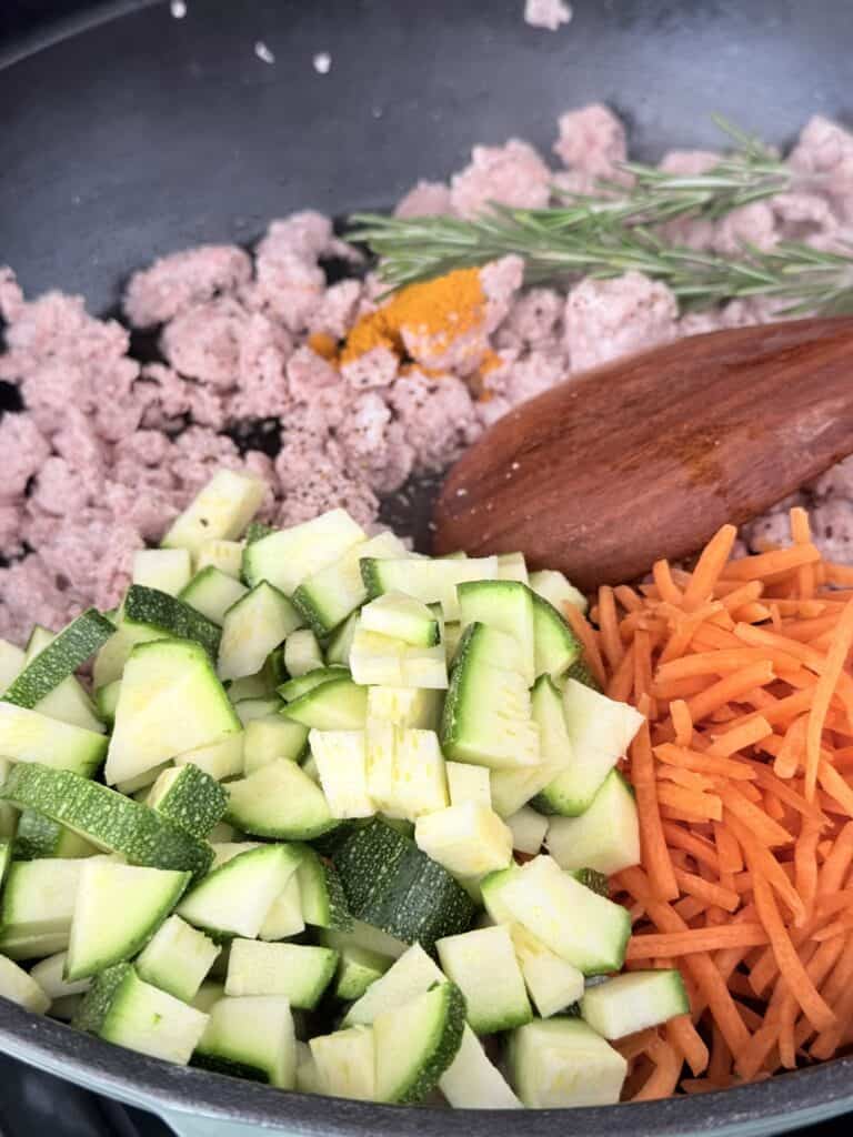 Ground beef brown in a pan with turmeric seasoning and seasoning, two sprigs of rosemary, shredded carrots, and zucchini in a pan on the stove top with a wooden spoon inside the pan.