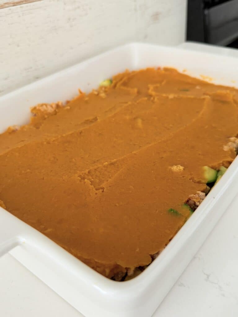 Ground meat and veggie mixture in a white baking dish with sweet potato mash on the top, all on the counter, ready to be baked.