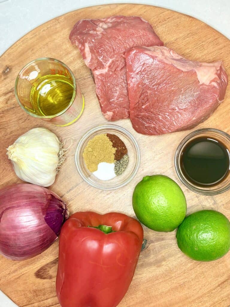 steak, bell pepper, limes, onion, seasoning, oil, garlic, and Worcestershire sauce on a wooden board