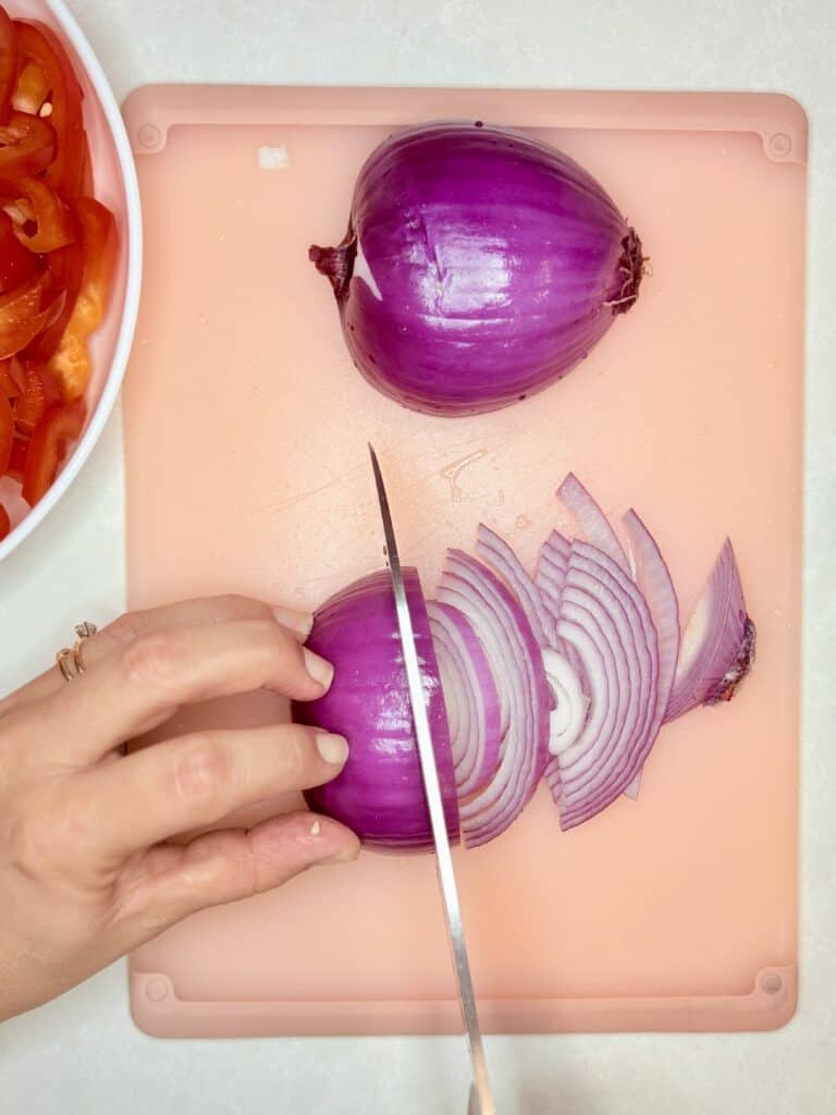 a hand holding a red onion being thinly sliced with a chef's knife on a cutting board