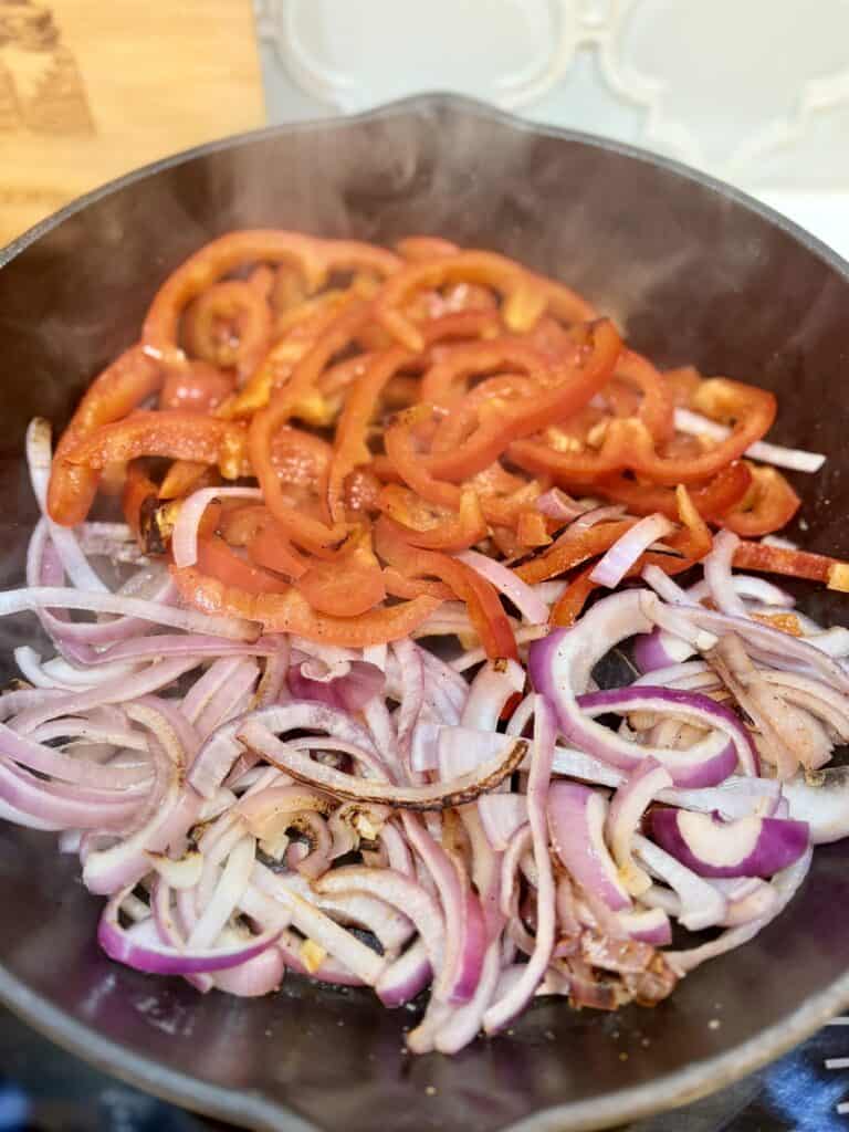 sliced red onions and bell peppers browning in a cast iron skillet on a stove