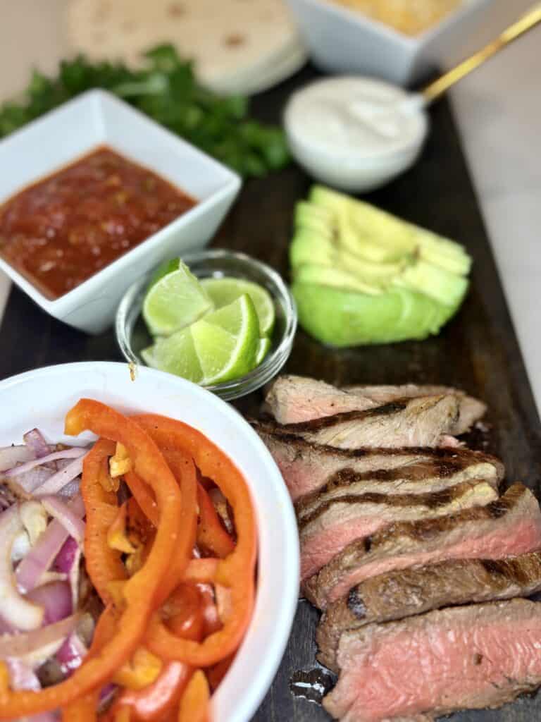 a cutting board with grilled steak slices, cooked red peppers and onions, sliced avocado, sliced limes and other fajita toppings