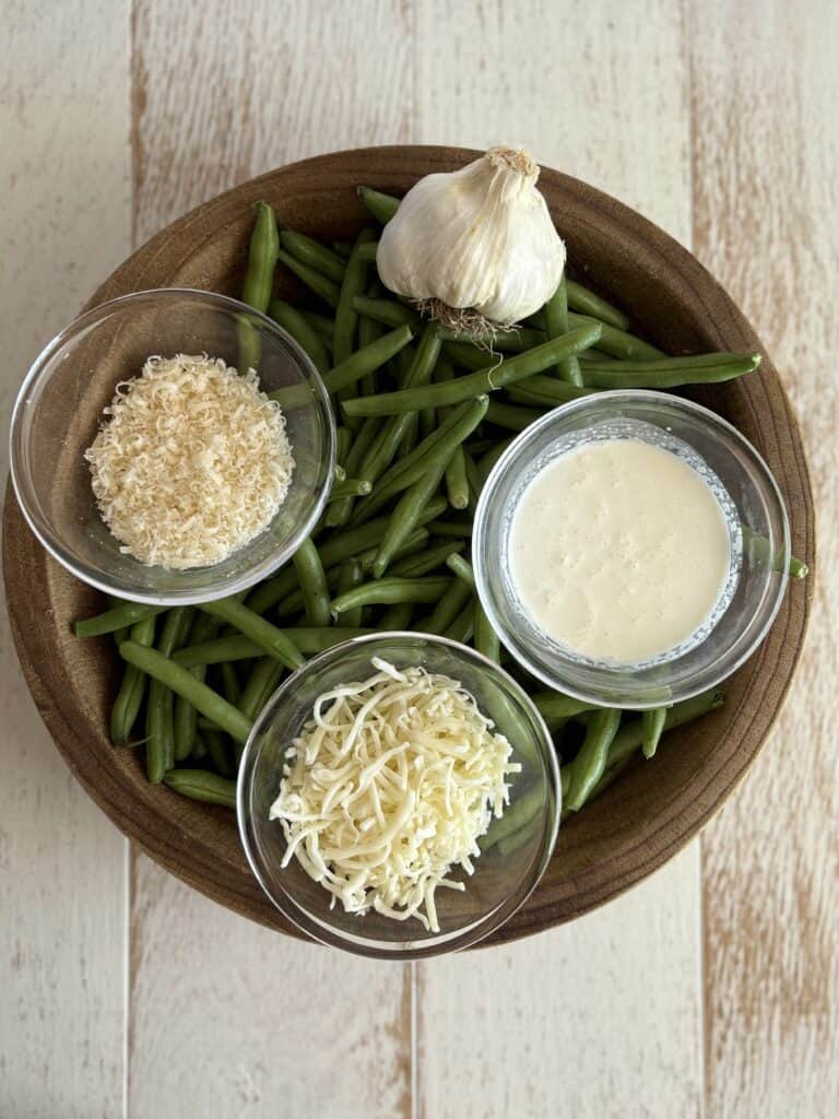 fresh green beans in a wooden bowl with small bowls of parmesan and mozzarella cheese, heavy cream and a bulb of garlic