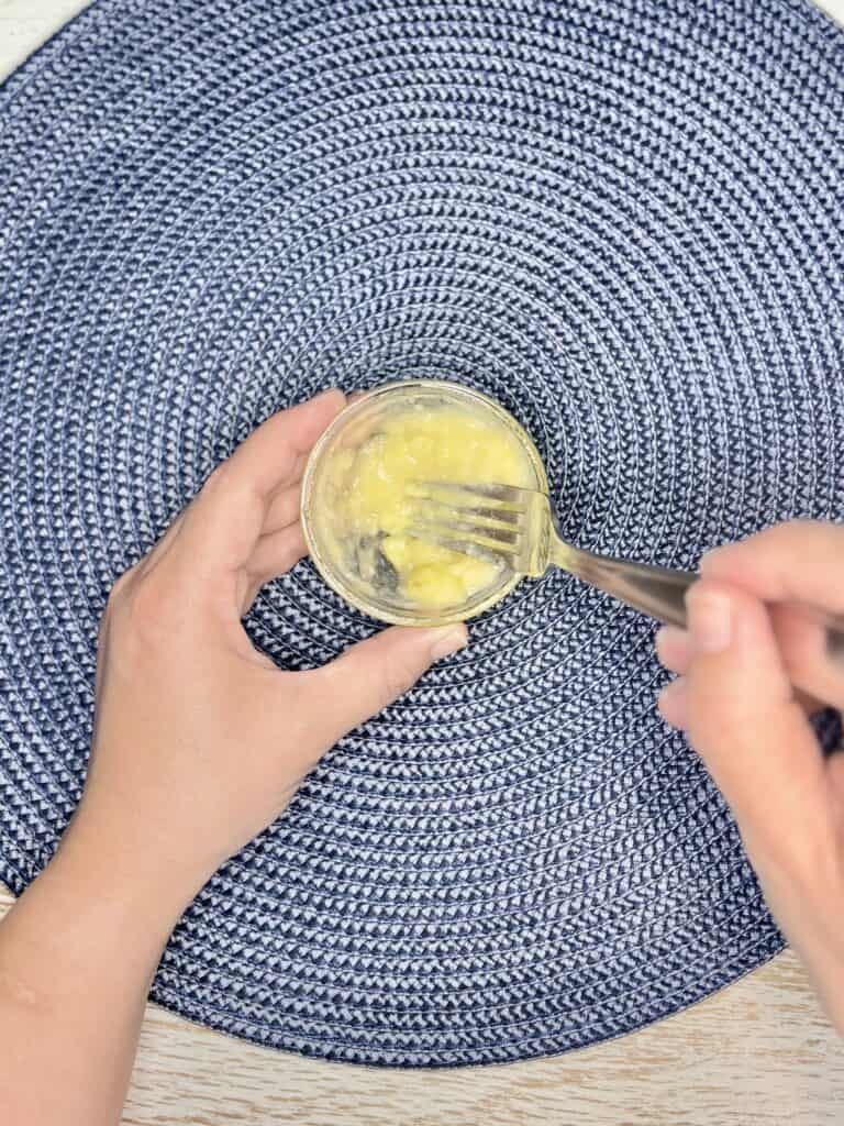 a hand using a fork to mash banana slices in a bowl