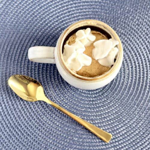 a mug with a muffin cooked in side topped with whipped cream next to a spoon