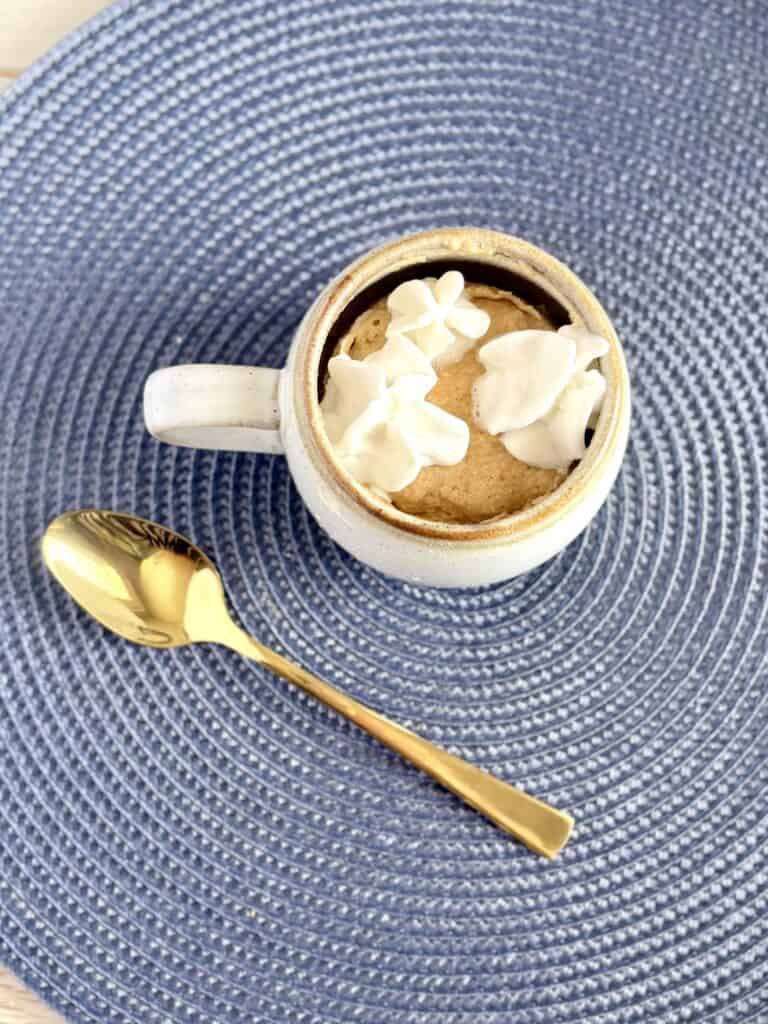 a mug with a muffin cooked in side topped with whipped cream next to a spoon