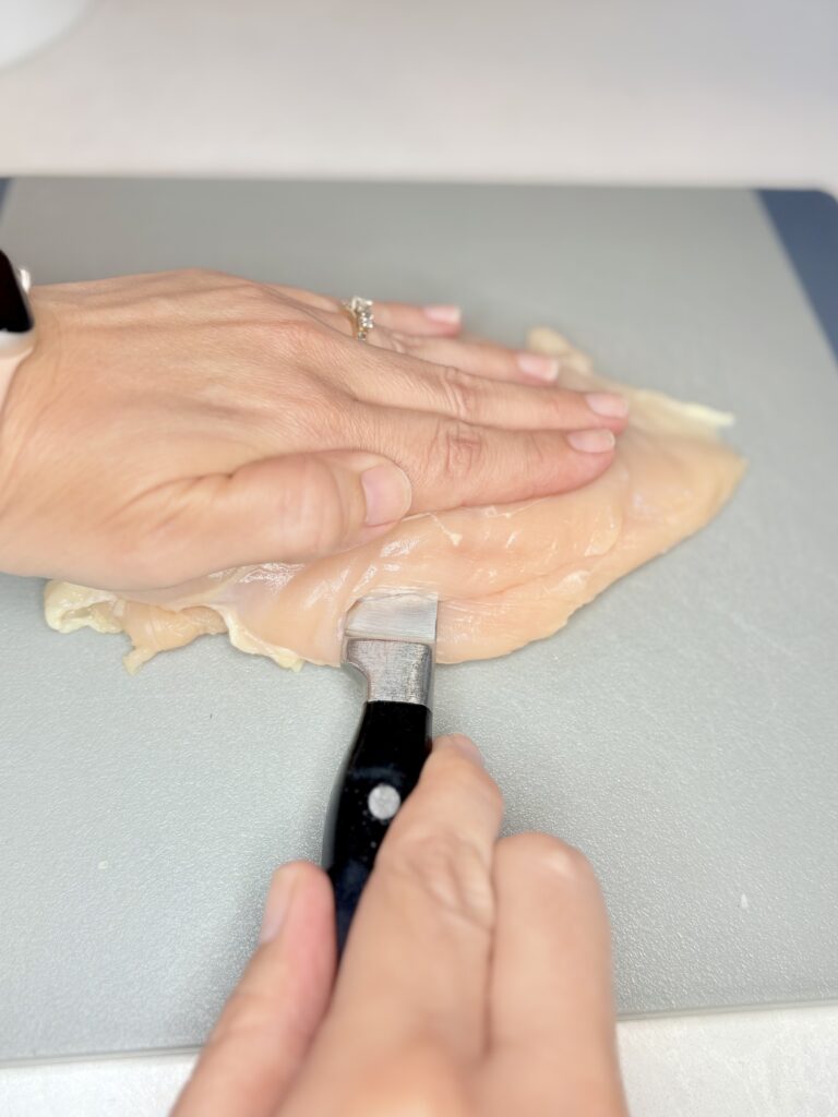 a hand using a small knife to slice a chicken breast into two thinner pieces