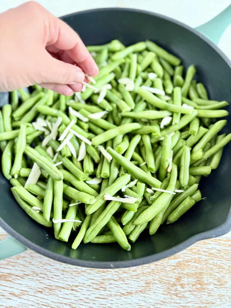 a hand sprinkling sliced garlic over green beans in a skillet