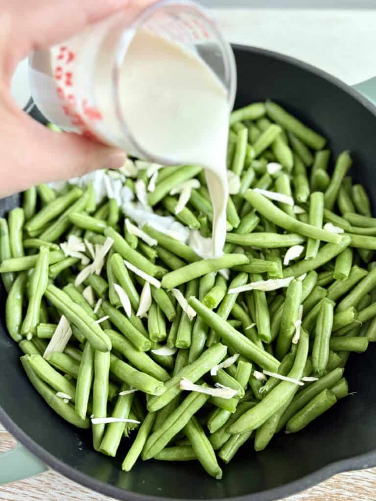 a hand pouring cream over green beans in a skillet