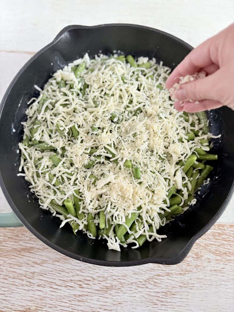 a hand sprinkling grated cheese over green beans in a skillet