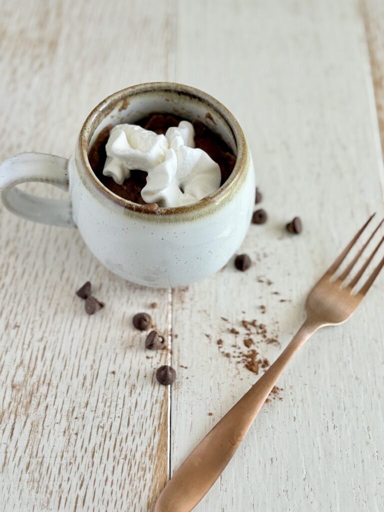 a muffin in a mug next to a fork with chocolate chips sprinkled around