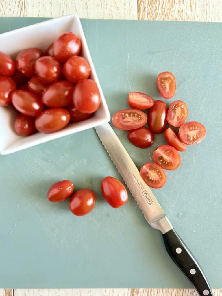 a knife slicing grape tomatoes in half next to a bowl of grape tomatoes