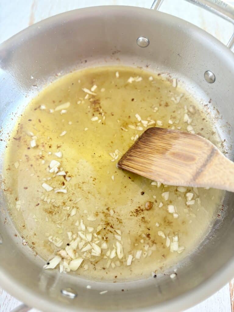 s wooden spatula scraping the bottom of a skillet containing garlic, wine and broth