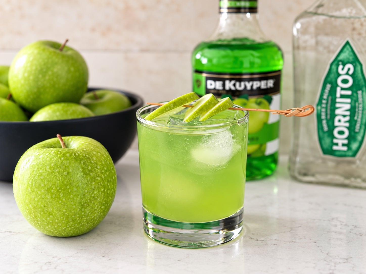 Bright green apple margarita with lime wedge garnish on a glass, surrounded by green apples and bottles of De Kuyper and Hornitos in the background.