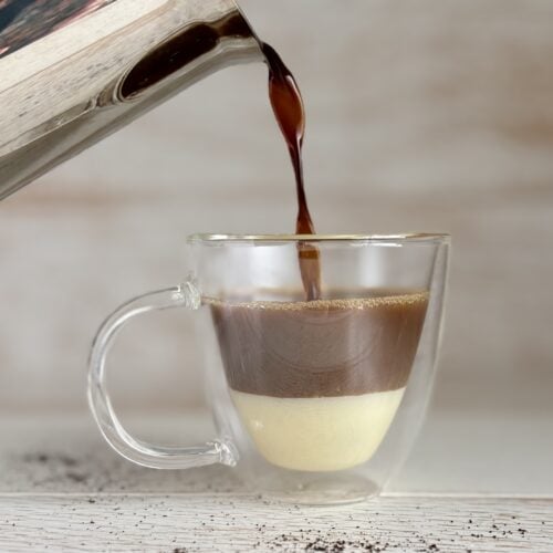 A layered Café Bombón coffee in a clear glass cup with espresso being poured from a metal pot.