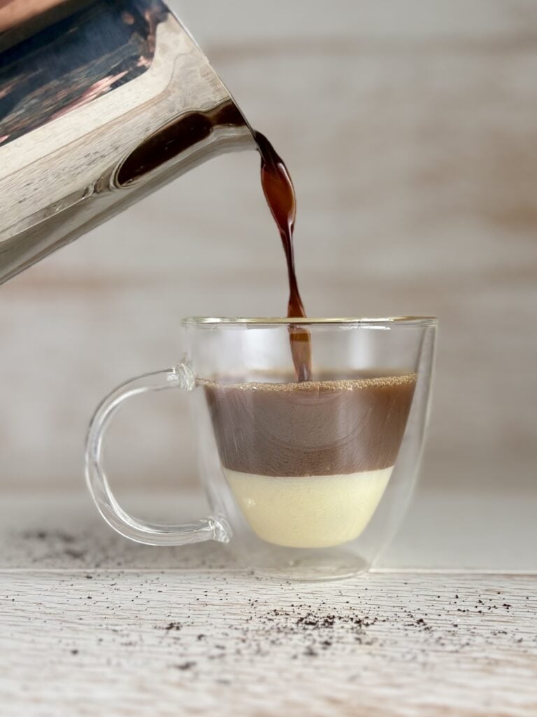A layered Café Bombón coffee in a clear glass cup with espresso being poured from a metal pot.