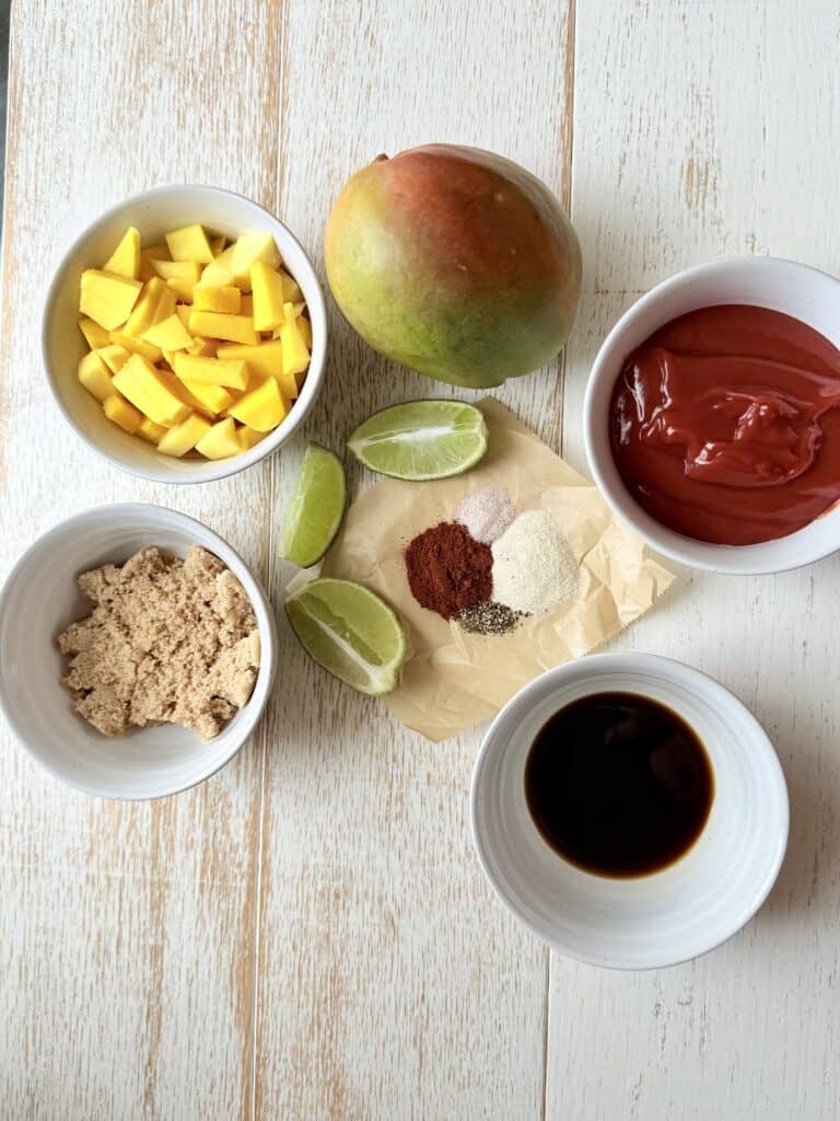 A ripe mango, diced mango in a small white bowl, brown sugar in a small white bowl, ketchup in a small white bowl, soy sauce in a small white bowl, with parchment paper, lime wedges, garlic powder, salt, and pepper, and smoked paprika all on the counter.