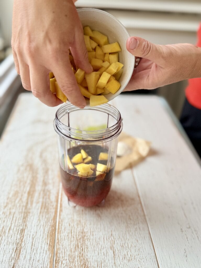 Adding mango pieces to a plastice high-speed blender cup on the counter.