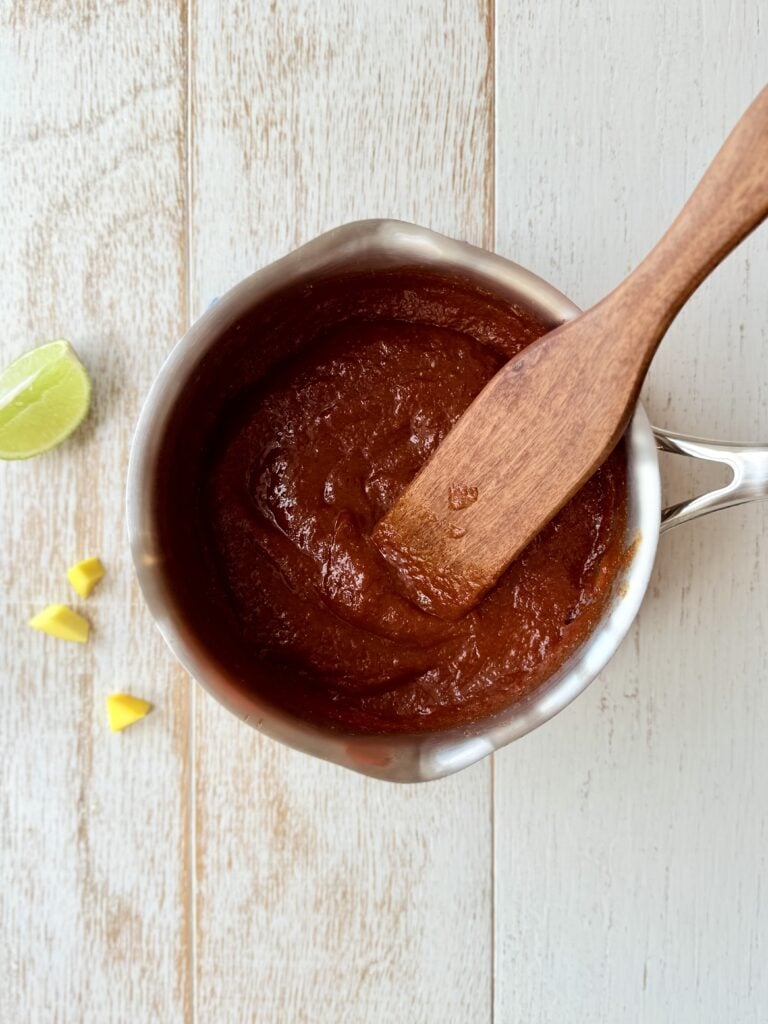 Mango BBQ sauce in a saucepan with a wooden spoon, on the counter. with a wedge of lime and mango pieces.
