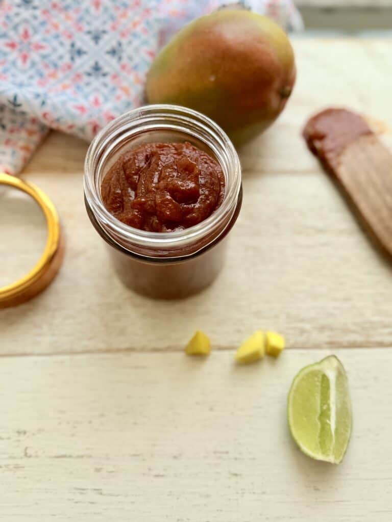Mango BBQ sauce in a mason jar on the counter. Surrounded by a ripe mango, mango pieces, a wooden spoon with BBQ sauce on it, and a blue and red hand towel in the background.