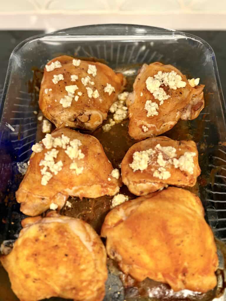 blue cheese crumbles sprinkled on to cooked chicken thighs in a glass baking dish