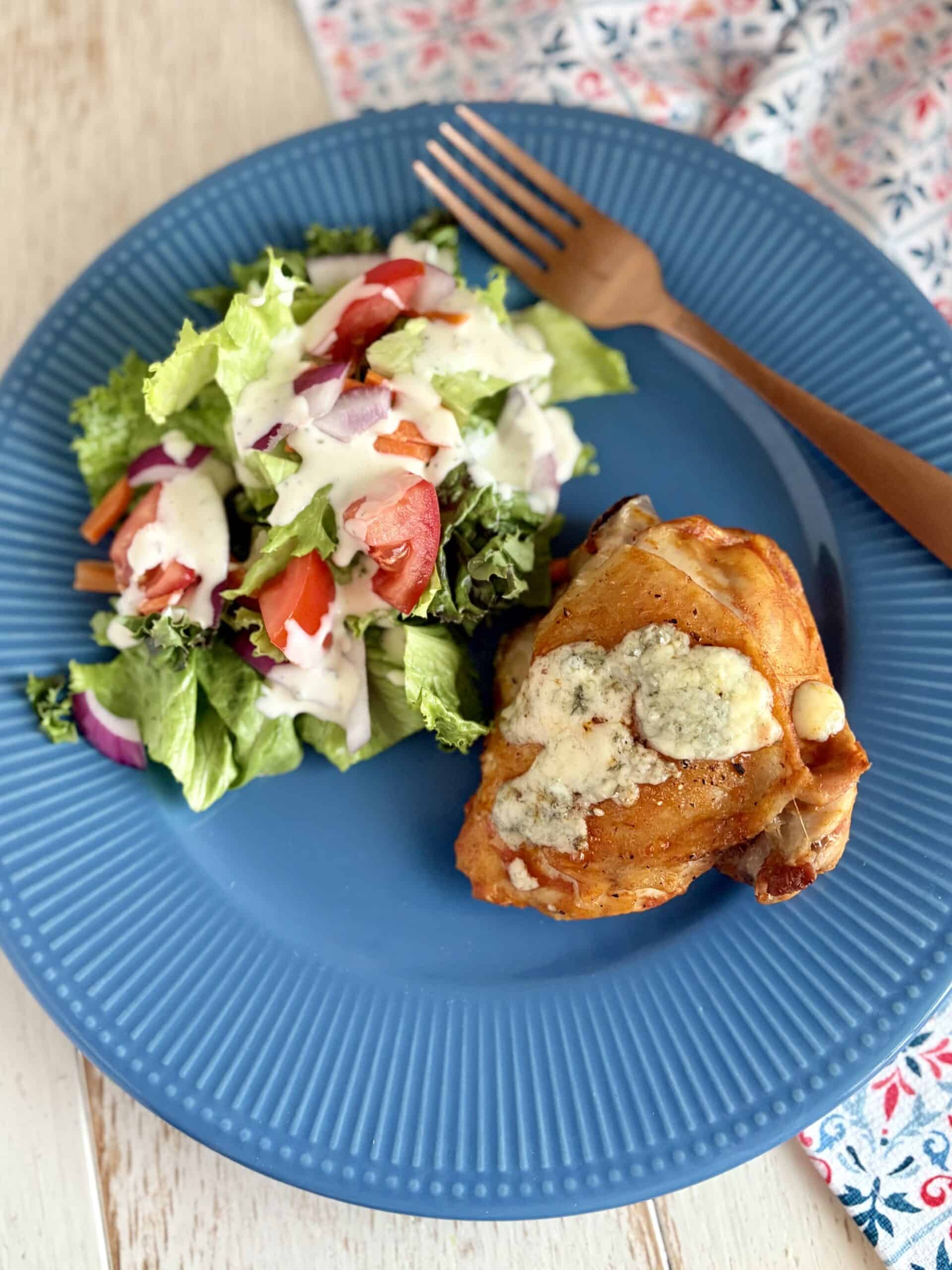 Baked Buffalo chicken thighs next to a salad with blue cheese dressing.