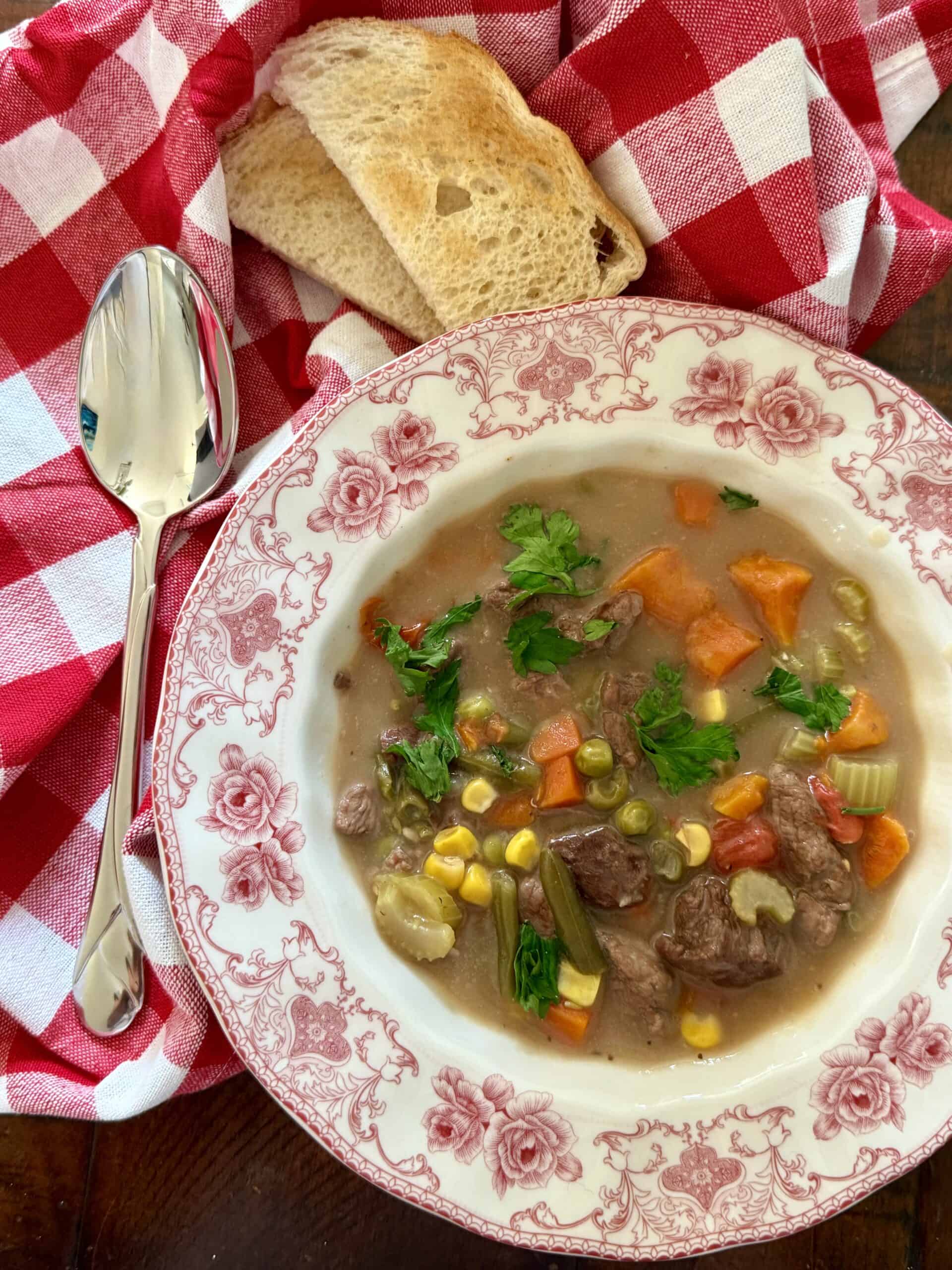 Steak stew with red wine sauce in a red and white bowl on a dark wood table with a red and white checker hand tool around it with toast and a silver spoon.