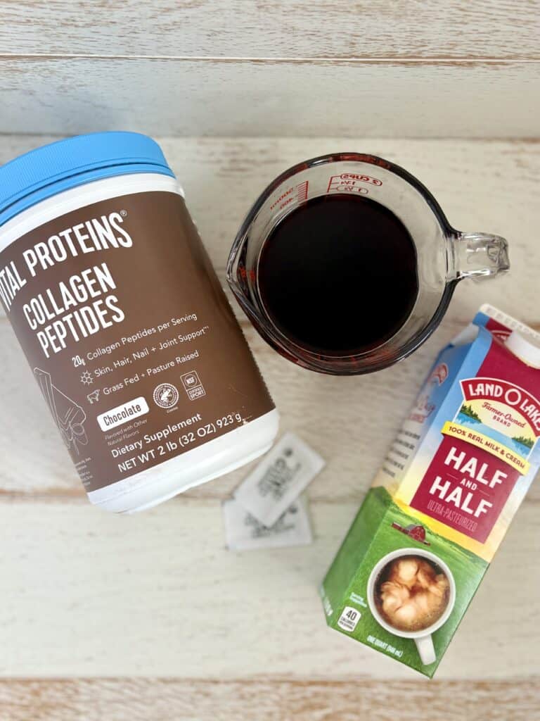 protein powder, half and half, a measuring cup with coffee in it and packets of sweetener on a wooden board