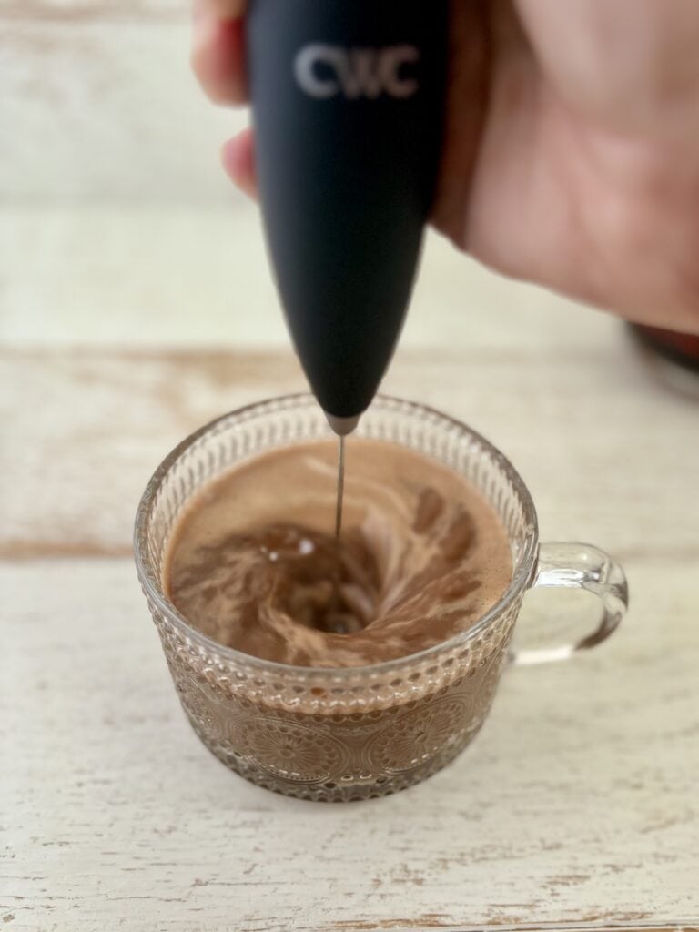 a frother being used to blend coffee and protein powder together in a mug