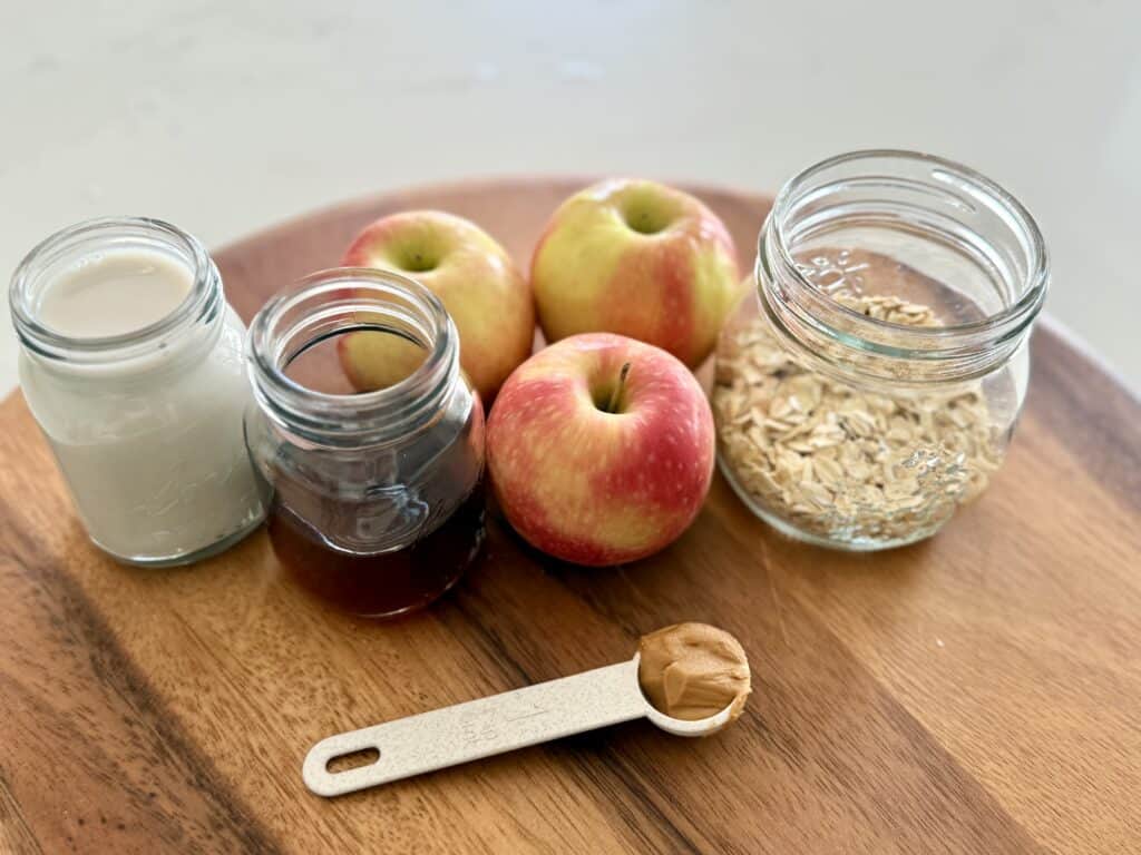 vanilla unsweetened almond milk in a small glass jar, maple syrup in a glass jar, oats in a glass jar with three small apples and a teaspoon of peanut butter in a measuring spoon, all on a brown cutting board on the white countertop.