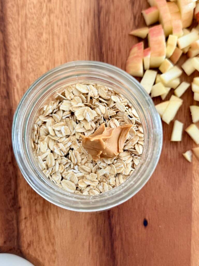 oats and peanut buttter in a glass jar on a brown cutting board with diced apple pieces on it.