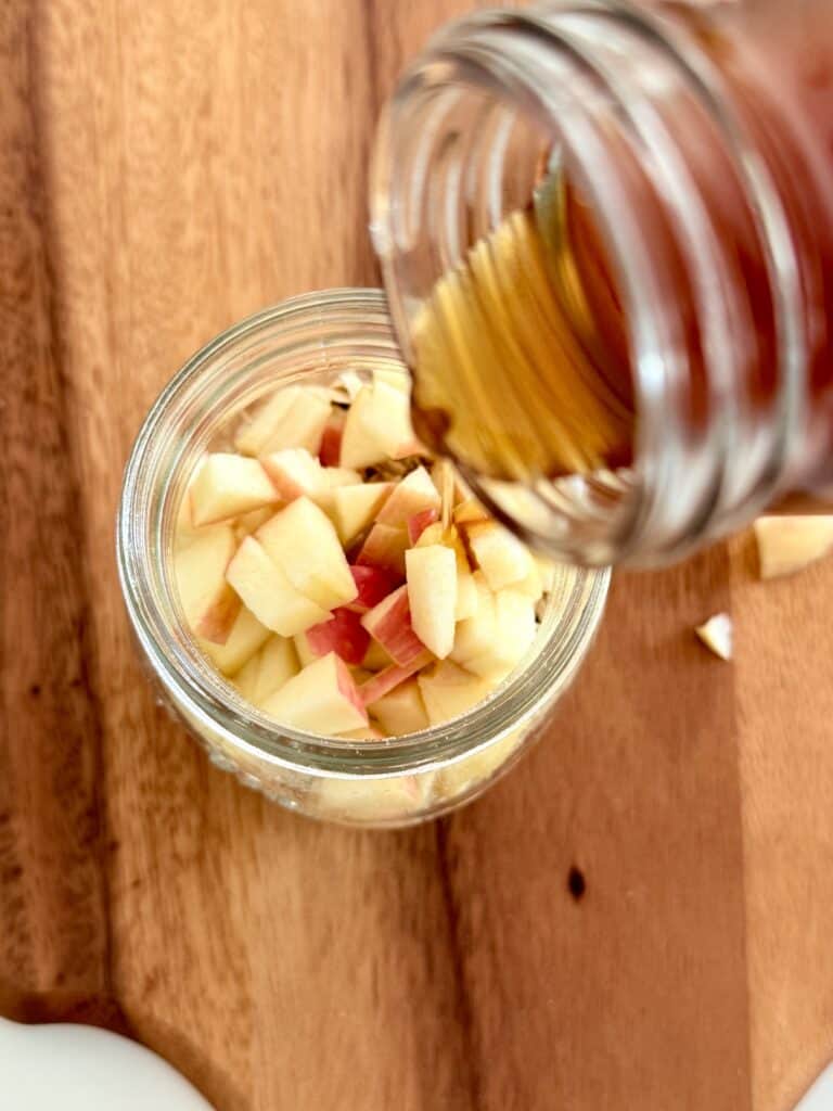 pouring maple syrup into a glass jar filled with oats, almond milk, and diced apple pieces on a brown cutting board.