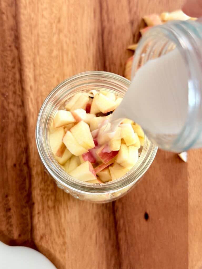 pouring vanilla unsweetened milk into a glass jar with oats and diced apple pieces in it on a brown cutting board on the counter.
