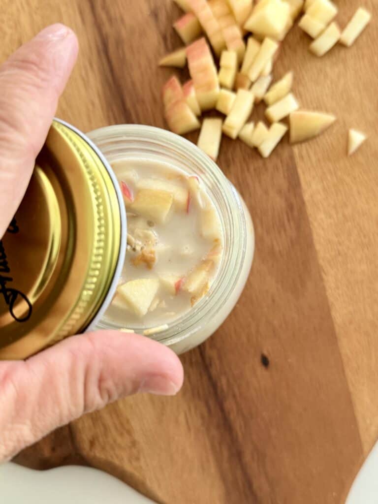 Putting a lid on a glass jar filled with the apple peanut butter overnight oats on a brown cutting board with diced apple pieces above.