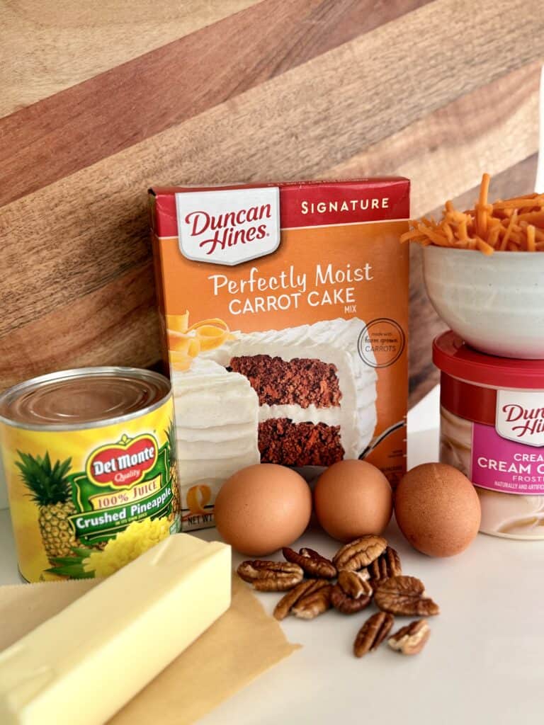 1 can of cream cheese frosting, a can of crushed pineapple, three eggs, a stick of butter, pecans, shedded carrots in a white bowl and one box of carrot cake mix.