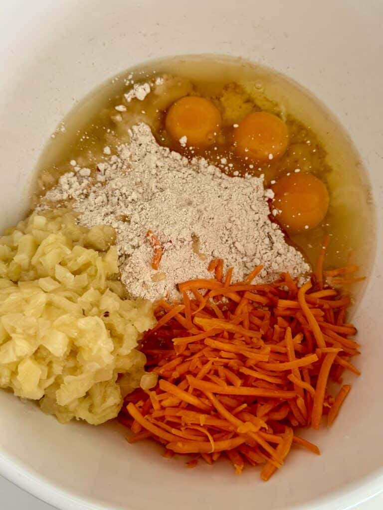 Cracked eggs, cake mix, shredded carrots, and crushed pineapple in a large white bowl on the counter.