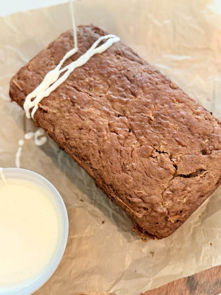 Warmed cream cheese frosting is being drizzled with a spoon onto the carrot cake loaf on parchment paper on the counter.