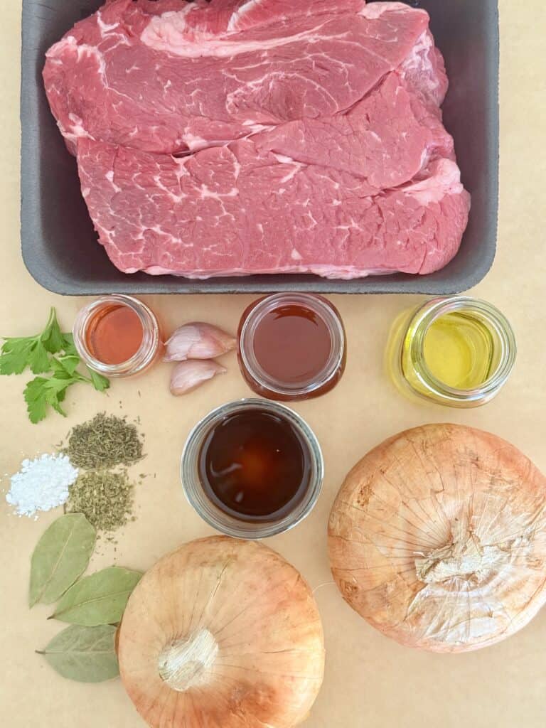 Chuck roast, dried oregano, dried thyme, cornstarch, olive oil oil in a glass jar, two yellow onions, worcestershire sauce in a glass jar, two garlic cloves, low sodium beef broth in a glass jar, red wine, glass jar, three whole leaves and fresh parsley, all impressing paper on the counter.