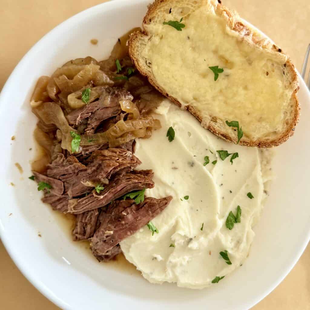 Easy Savory onion pot roast on a white plate with mashed potatoes and cheesy bread all on brown parchment paper on the counter.