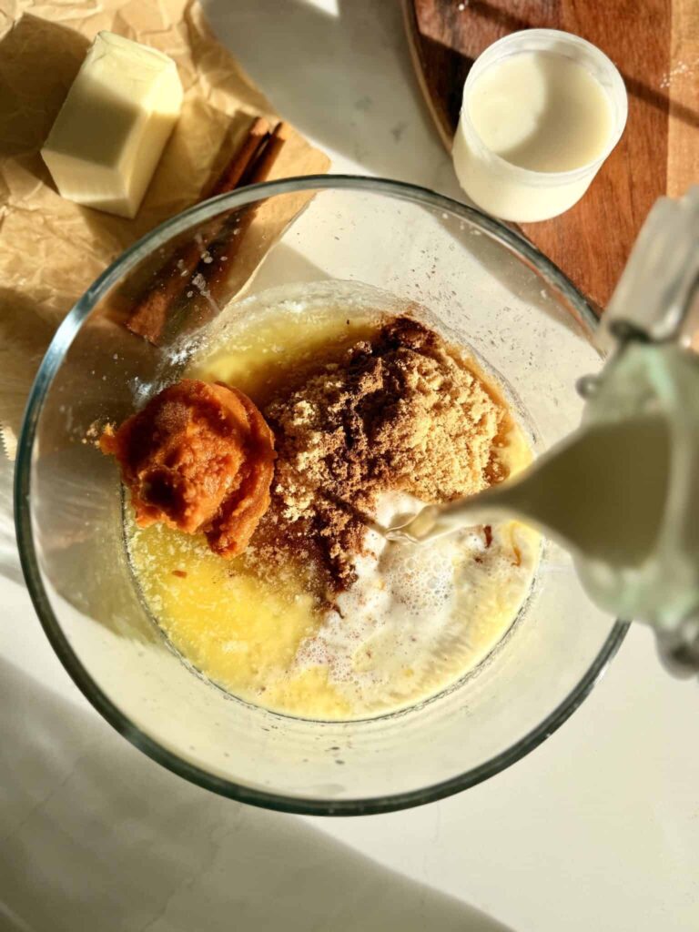 Pouring heavy cream into a clear bowl with melted butter, sugar, and canned pumpkin all on the counter.