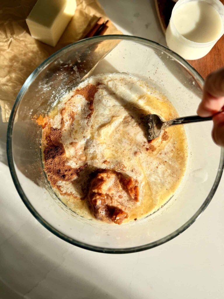 Mixing together melted butter, sugar, canned pumpkin, and heavy cream altogether in a glass bowl on the counter