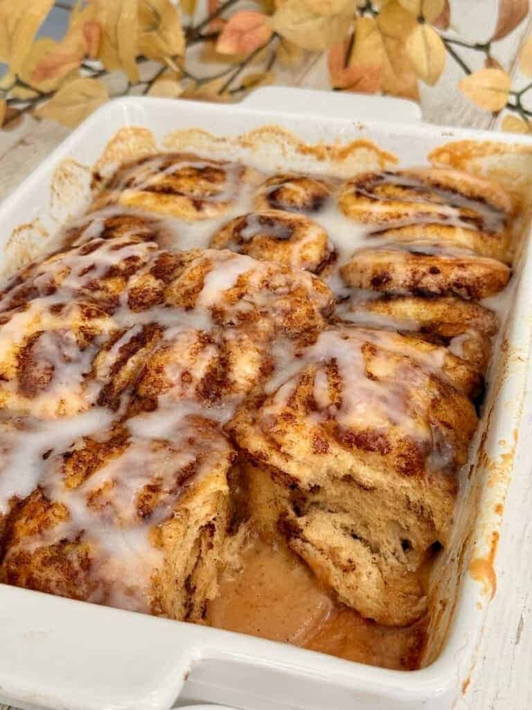 Pumpkin cinnamon rolls with icing in a baking dish, topped with autumn leaves for seasonal presentation.