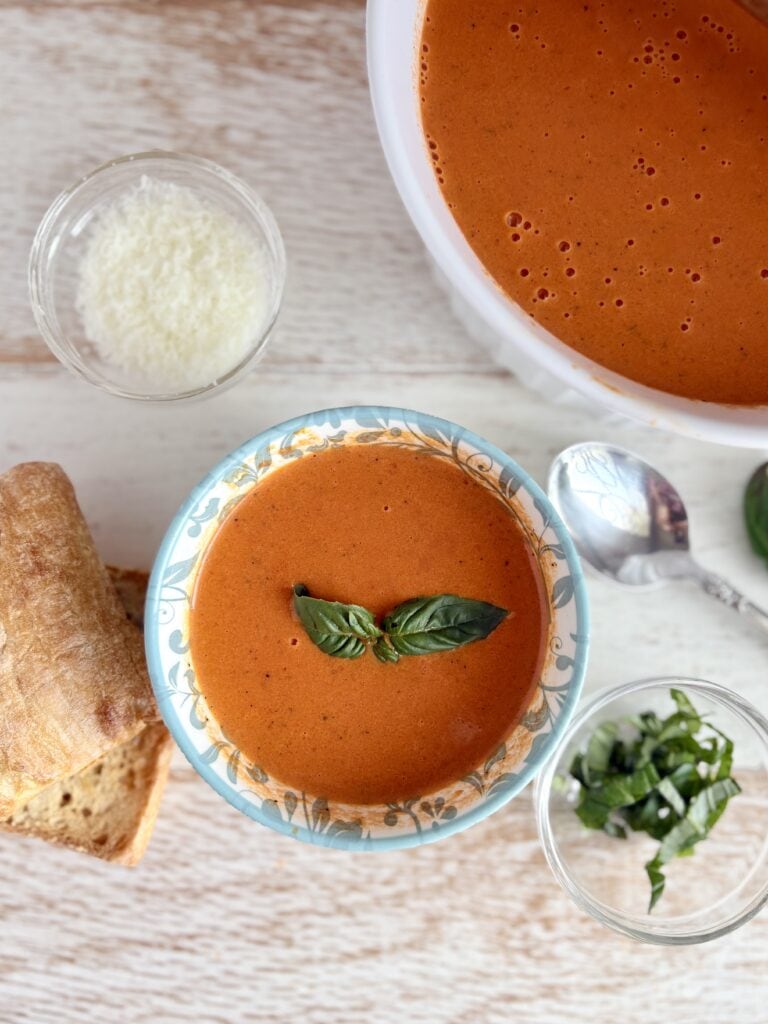 Creamy roasted red pepper soup topped with fresh basil leaves, served with toasted bread, grated cheese, and garnished with chopped green herbs; perfect for a comforting meal.