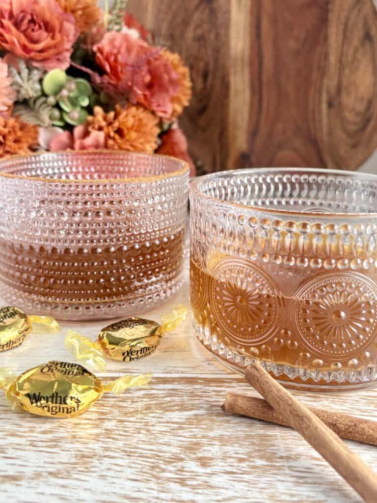 2 glass mugs containing spiked caramel cider with cinnamon sticks and caramel candies