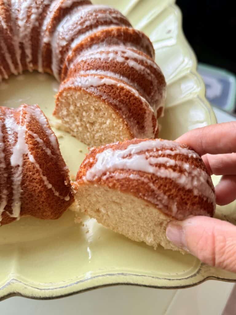 Bisquick Bundt cake on a green cake platter on the counter, with a hand grabing a sliced piece of cake.